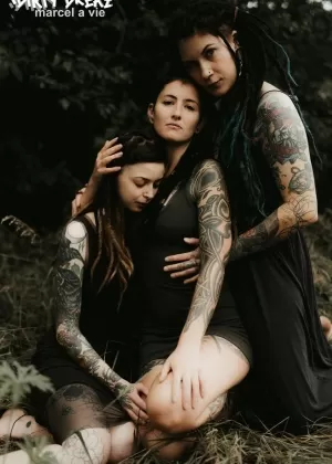 Три татуированные девушки обмениваются лесбийскими поцелуями в высокой траве возле ели